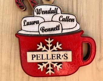 Può includere: Una tazza di legno rossa con un fiocco di neve bianco e il testo "PELLER'S" sul davanti. La tazza è piena di marshmallow bianchi con i nomi "Wendall", "Callen", "Laura" e "Bennett" scritti su di essi. Un bastoncino di zucchero rosso e bianco è sopra i marshmallow.
