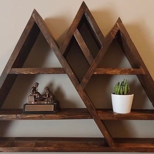 May include: A wooden mountain-shaped shelf with multiple shelves. The shelf is stained a dark brown color and has a rustic look. There is a small figurine on one shelf and a potted succulent on another.
