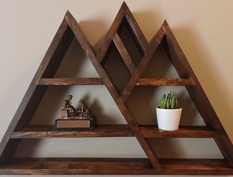 May include: A wooden mountain-shaped shelf with multiple shelves. The shelf is stained a dark brown color and has a rustic look. There is a small figurine on one shelf and a potted succulent on another.