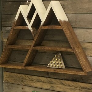 May include: A wooden mountain-shaped shelf with white painted accents. The shelf has five shelves and is mounted on a wood plank wall. A small photo of a child is displayed on a shelf below the mountain shelf.