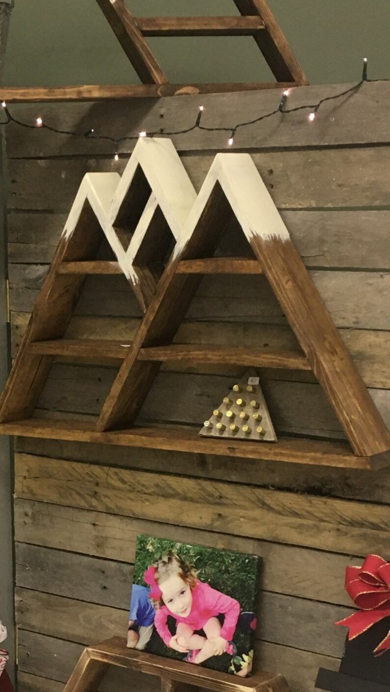 May include: A wooden mountain-shaped shelf with white painted accents. The shelf has five shelves and is mounted on a wood plank wall. A small photo of a child is displayed on a shelf below the mountain shelf.