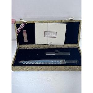 May include: A decorative sword with a patterned blade rests in a rectangular box lined with black velvet and featuring a gold-toned exterior with intricate patterns. The box includes a white card with red text and a small black stand.