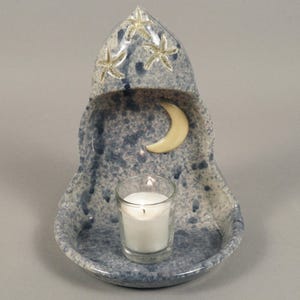 May include: A ceramic candle holder with a blue and white speckled glaze, featuring a pointed arch shape. The interior displays a cream-colored crescent moon, while the exterior top is decorated with two starfish. A clear glass votive candle holder sits at the base.