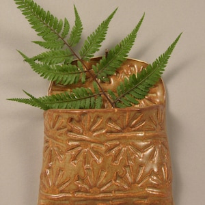 May include: A wall-mounted ceramic pocket planter with a glossy tan glaze and a repeating embossed sunburst pattern. The pocket holds a single green fern frond with delicate, feathery leaves extending outward from the top opening of the vessel.