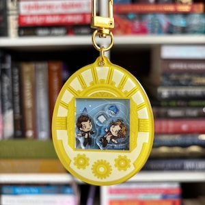 Può includere: Un ciondolo in acrilico giallo con chiusura color oro. Il ciondolo presenta un design con due personaggi dei cartoni animati all'interno di una cornice, con sfondo blu e piccole illustrazioni di libri. Il ciondolo ha un design a forma di sole e dettagli a ingranaggi.