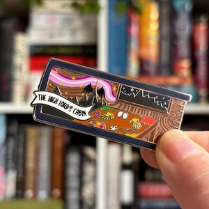 Pode incluir: Pin esmaltado com uma cena de cabana e o texto "THE HIGH LORD'S CABIN". O pin tem uma borda azul escura e retrata o interior de uma cabana com uma cordilheira e um céu colorido. O design inclui uma paleta e pincéis.