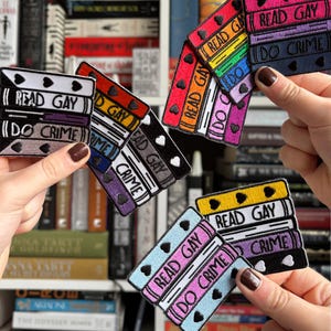 以下が含まれることがあります： 「READ GAY」と「DO CRIME」の文字が書かれた、本が積み重ねられた刺繍パッチ。パッチは黒、赤、青、黄色など様々な色があり、ハートのアクセントが付いています。背景には本棚が写っています。