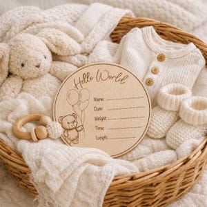 May include: A wooden baby announcement sign with the words "Hello World" and space for details. The sign is in a wicker basket with a stuffed bunny, baby clothes, and a wooden rattle. The sign has a teddy bear illustration.