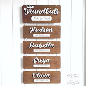 May include: Wooden sign with the text "GREAT Grandkids Make Life Grand" and a list of names with dates, including Hudson, Isabella, Freya, and Olivia. The sign is brown with white lettering and is hanging on a white wall.