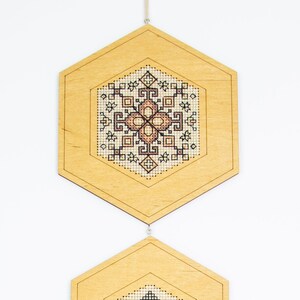 May include: Three wooden hexagonal ornaments with intricate geometric patterns in black and brown thread. The ornaments are suspended from a string and hang in a row.
