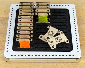 May include: A white square organiser tray with a black foam insert featuring horizontal slots. The tray holds multiple light-coloured wooden thread bobbins, some wrapped with colourful embroidery floss, and two decorative wooden spools with floral patterns.