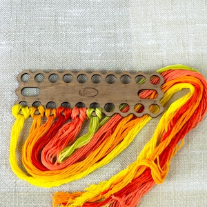May include: A wooden thread organizer with multiple holes, holding colorful embroidery floss in shades of yellow, orange, and green. The floss is knotted and arranged, creating a vibrant display against a neutral background. The organizer has the text "La Casita" on it.