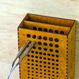 May include: A wooden knitting needle gauge and organizer, stained in a warm yellow hue. The organizer features multiple holes for needle sizes, with two needles inserted. The gauge is marked with numbers and measurements in inches.