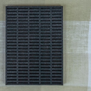 May include: A dark gray, rectangular mat with evenly spaced, horizontal slots. The mat is made of a textured material and is displayed against a neutral background. The design suggests a focus on functionality and possibly drainage or ventilation.