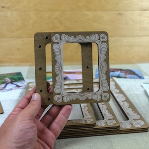 May include: Wooden photo frames with a decorative floral design. The frames are a light brown color with a lighter, off-white floral pattern. Several frames are stacked, and one is being held up, showing the design.