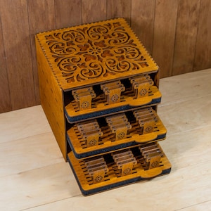 May include: A wooden storage box with three pull-out trays. The box and trays are a light brown color with intricate floral carvings on the top and tray edges. Each tray holds several small, rectangular items.