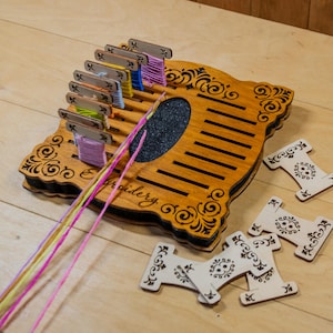 May include: Wooden embroidery floss organizer with a decorative design, holding spools of colorful thread. The organizer has a dark brown finish with a black center and the word "Embroidery" on the side. Several wooden bobbins are scattered around the organizer.