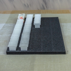 May include: Three stacks of white plastic containers with a rectangular black foam base. The containers are arranged on a beige and white checkered surface. The containers are of varying heights.