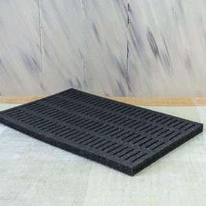 May include: A rectangular black foam insert with evenly spaced, parallel slits. The foam is placed on a light-colored, patterned surface. The background features a light-colored wooden wall.