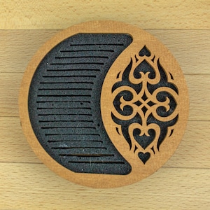 May include: A round wooden coaster with a crescent-shaped cutout filled with black material. The coaster features an intricate, symmetrical design on the right side, with heart and swirl motifs. The coaster is made of light brown wood.