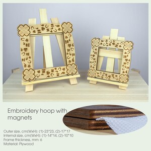 Op de afbeelding: Twee houten borduurringen met magneten, elk met een decoratief ontwerp van klaverbladeren en hoefijzers. De ringen staan op kleine houten ezelstandaards. De grotere ring meet 23 cm bij 23 cm en de kleinere 17 cm bij 17 cm. De tekst "Borduurring met magneten" is zichtbaar onder de afbeelding.