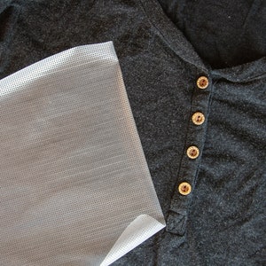May include: A close-up of a gray shirt with four brown buttons. A piece of clear, white, patterned fabric is lying on top of the shirt.