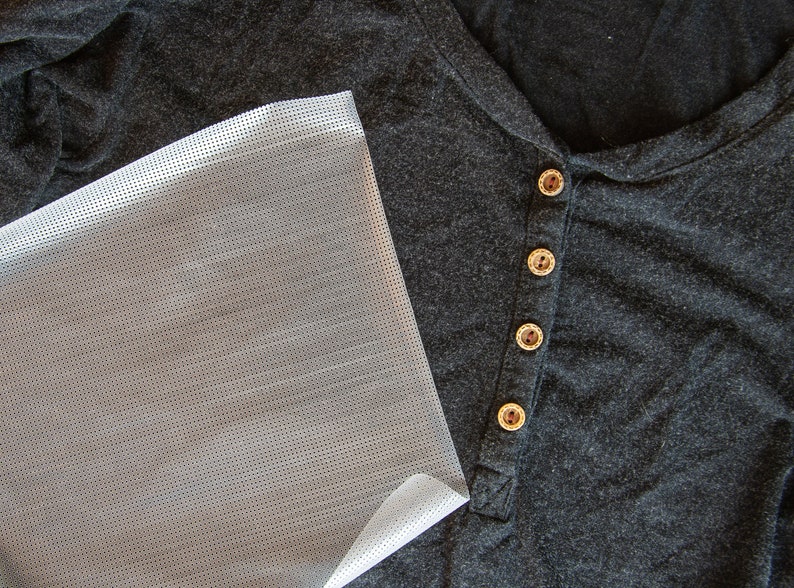 May include: A close-up of a gray shirt with four brown buttons. A piece of clear, white, patterned fabric is lying on top of the shirt.