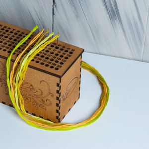 Peut inclure: Une boîte en bois avec des trous numérotés pour ranger du fil à broder. La boîte a un motif floral et est remplie de fil vert, jaune et orange.