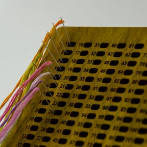 May include: A wooden needlepoint chart with multiple needles threaded with colorful embroidery floss. The chart has numbered holes for precise stitching. The floss colors include orange, green, and pink.