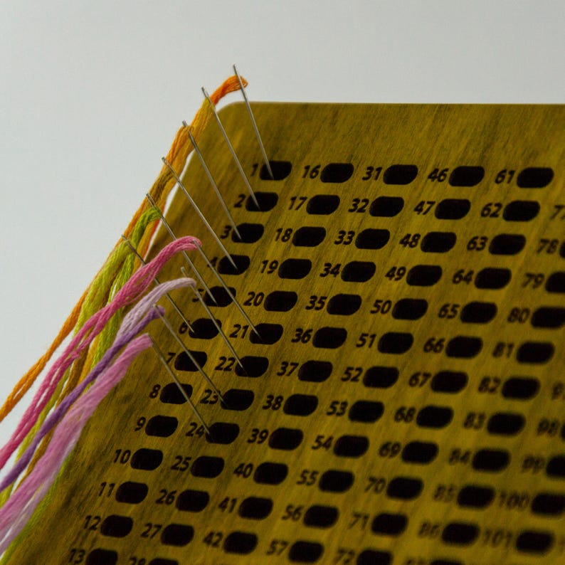 May include: A wooden needlepoint chart with multiple needles threaded with colorful embroidery floss. The chart has numbered holes for precise stitching. The floss colors include orange, green, and pink.