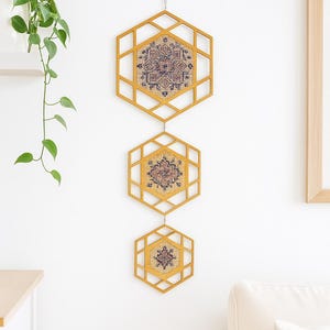 May include: Three geometric wall hangings with a honeycomb design. Each frame is a warm yellow color and holds a patterned textile with shades of blue, purple, and tan. The wall hangings are suspended from a metal hook.
