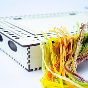 Pode incluir: Uma caixa de artesanato de madeira com tampa, com uma série de orifícios e ranhuras. Fios de bordar coloridos em tons de amarelo, laranja, verde e castanho derramam-se. Pequenas pedras preciosas coloridas estão espalhadas por perto.
