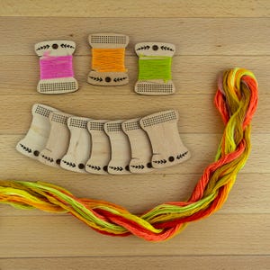 Peut inclure: Des bobines de fil à broder en bois et du fil à broder coloré. Trois bobines sont enroulées de fil rose, orange et vert. Sept bobines en bois supplémentaires sont disposées en dessous. Une longueur de fil multicolore est également présente.