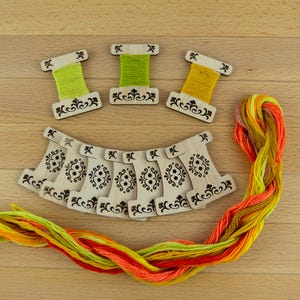 Peut inclure: Bobines en bois pour fil à broder avec des découpes décoratives, certaines contenant du fil vert et jaune. Une écheveau de fil à broder coloré dans les tons orange, jaune et vert est également présent. Les articles sont disposés sur une surface en bois clair.