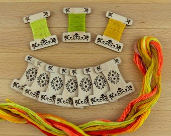 May include: Wooden embroidery floss bobbins with decorative cutouts, some holding green and yellow thread. A skein of colorful embroidery floss in shades of orange, yellow, and green is also present. The items are arranged on a light wood surface.