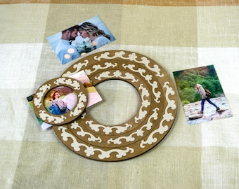 May include: Two circular wooden frames with a dark brown finish and light cream-coloured decorative scrollwork overlays. The larger frame features a wide circular opening, while the smaller frame holds a photograph of a person in a blue shirt.