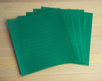 May include: A set of five square sheets of green plastic canvas, each featuring a uniform grid of small, evenly spaced holes. The sheets are arranged in a slightly fanned, overlapping stack, displaying a consistent vibrant green colour.