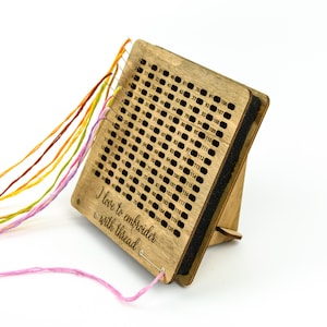 Op de afbeelding: Houten draadorganizer met een standaard, met een raster van genummerde gaten om borduurgaren op te bergen. De organizer heeft een zwarte schuimrug en de tekst "Ik hou van borduren met draad" is gegraveerd op de voorkant.