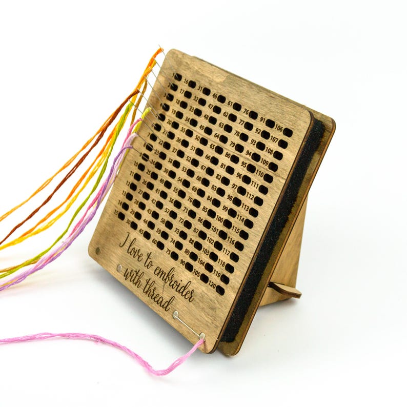Op de afbeelding: Houten draadorganizer met een standaard, met een raster van genummerde gaten om borduurgaren op te bergen. De organizer heeft een zwarte schuimrug en de tekst "Ik hou van borduren met draad" is gegraveerd op de voorkant.