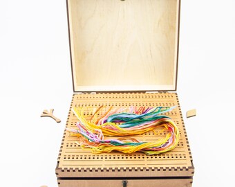 May include: A wooden box with a hinged lid and a latch. The box has a gridded interior with rows of holes for storing embroidery floss. A bundle of colorful embroidery floss is inside the box.
