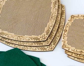 May include: A stack of laser-cut wooden cross-stitch canvases with decorative floral and leaf border designs. The canvases are light brown with numerous small holes for stitching. A piece of dark green felt is in the foreground.