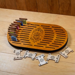 Pode incluir: Um organizador de linha de bordar em madeira de formato oval com ranhuras horizontais e um motivo floral central gravado a laser. Várias bobinas de madeira enroladas com linha colorida estão inseridas nas ranhuras, com cinco bobinas vazias à frente.