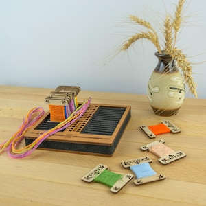 May include: A wooden weaving loom with colorful threads and spools. The loom is rectangular with a dark gray interior and a brown wooden frame. Several spools of thread in various colors are scattered around the loom, and a vase with wheat stalks is in the background.