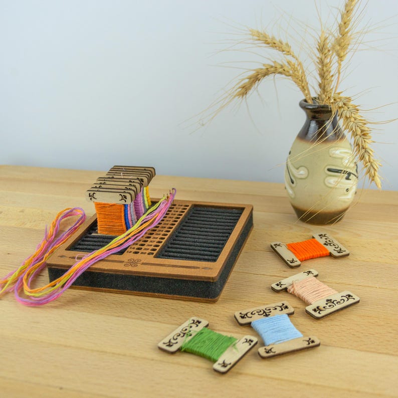 May include: A wooden weaving loom with colorful threads and spools. The loom is rectangular with a dark gray interior and a brown wooden frame. Several spools of thread in various colors are scattered around the loom, and a vase with wheat stalks is in the background.