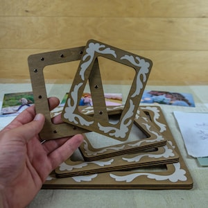 May include: Wooden photo frames in various sizes. The frames are light brown with white decorative accents. The frames are square with rounded corners and small holes for attaching photos. Some photos are visible in the background.