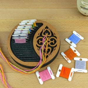 May include: A wooden embroidery floss organizer with a decorative cutout design, holding spools of colorful thread. Several loose spools of thread in blue, orange, and pink are scattered nearby. The threads are in shades of pink, yellow, and orange.