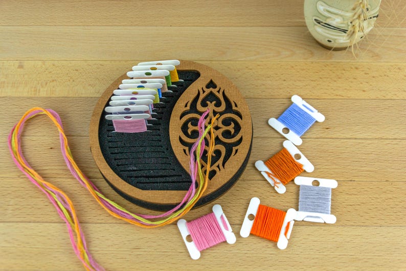 May include: A wooden embroidery floss organizer with a decorative cutout design, holding spools of colorful thread. Several loose spools of thread in blue, orange, and pink are scattered nearby. The threads are in shades of pink, yellow, and orange.