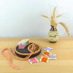 May include: A wooden embroidery floss organizer with a black interior and a decorative cutout design. Spools of colorful embroidery floss are arranged around the organizer. A small vase with wheat stalks is in the background.