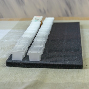 May include: Two stacks of white plastic thread bobbins are arranged on a dark grey foam pad. The bobbins are neatly aligned, showcasing their intricate design. The foam pad provides a contrasting backdrop, highlighting the bobbins' details.