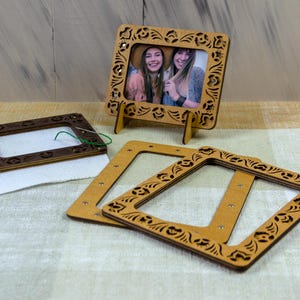 May include: Wooden photo frames with decorative floral cutouts. One frame holds a photo of two smiling people. Another frame is on a white cloth with a needle and green thread. Two additional frames are also visible.